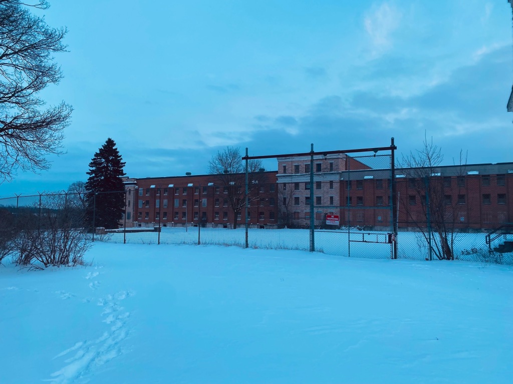 Photo of D-Cell Block prisoner wing. Four story stone and brick structure is behind a barbed wire chain link fence with a no-trespassing sign on the gate. There are footprints in the snow at this popular dog walking spot. The sky is overcast and dusk is near. There are few barren trees and bushes, and one evergreen. 