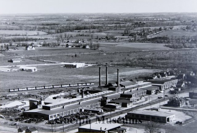 A black and white aerial view of the International Malleable Iron Company factory in 1948. The large factory has 3 smoke stacks. A train is parked in the background. Beyond the manufacturing site is a few homes and farmland.